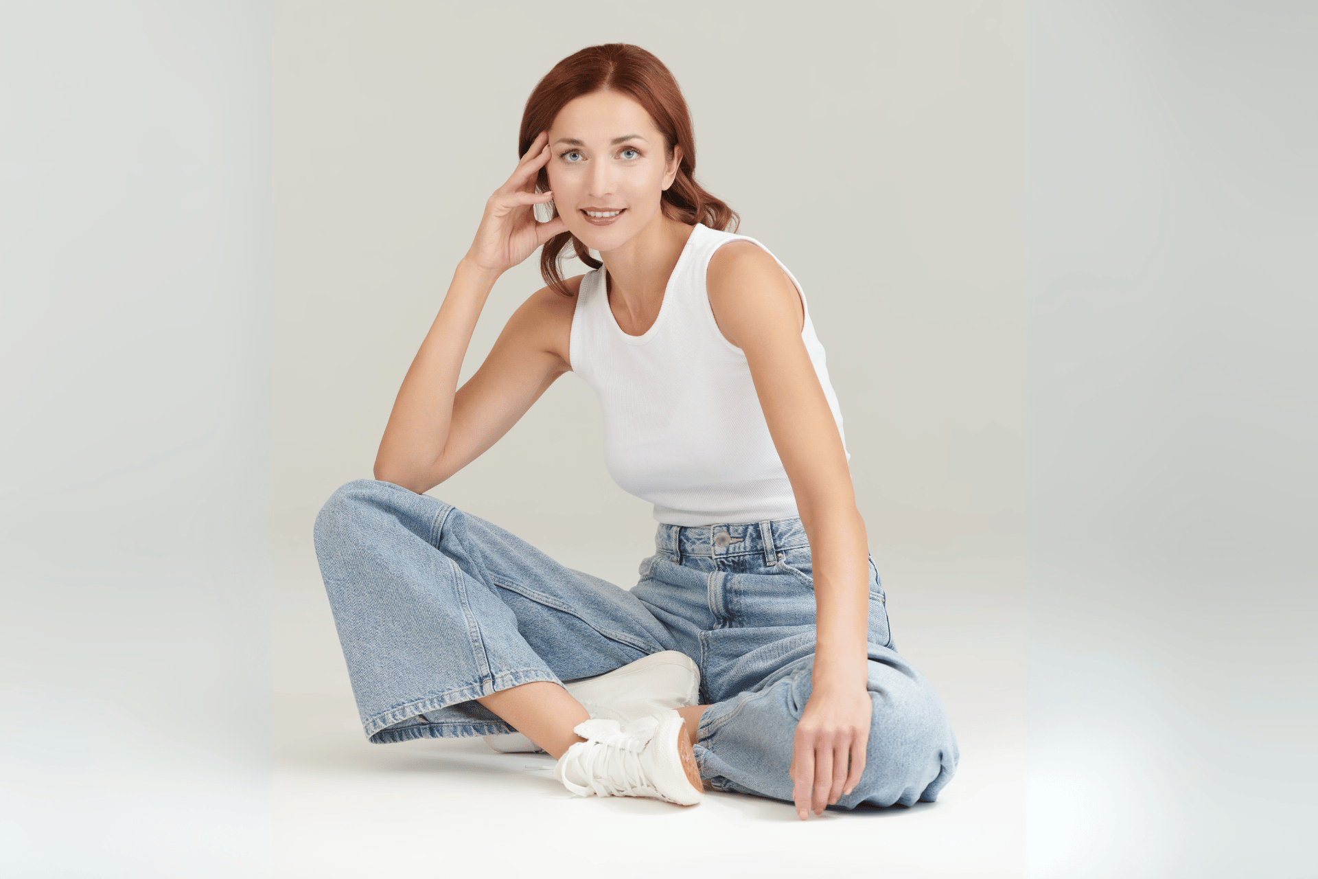 image of an attractive middle aged woman sitting in a studio on a grey background wearing denim jeans and a white t-shirt